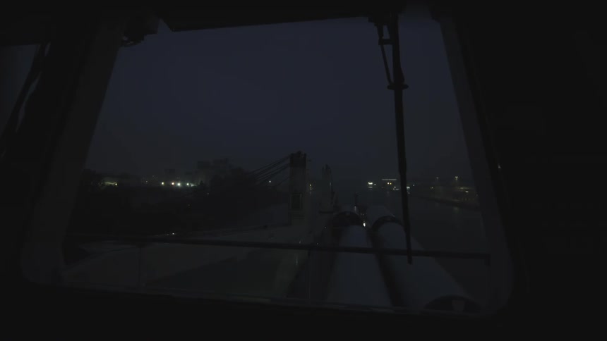 From the bridge of a cargo ship, large industrial components are transported through a misty canal before dawn