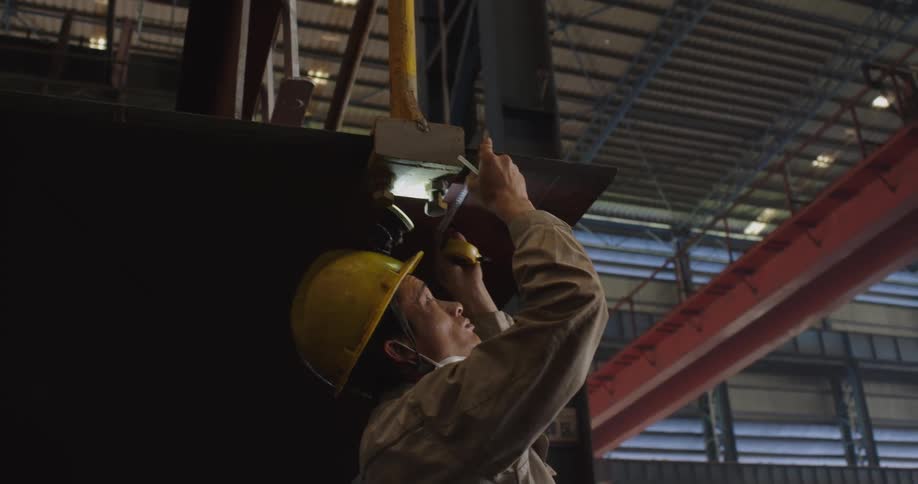 A focused shipyard worker in a hard hat meticulously inspects a large steel component suspended by a crane inside a vast factory