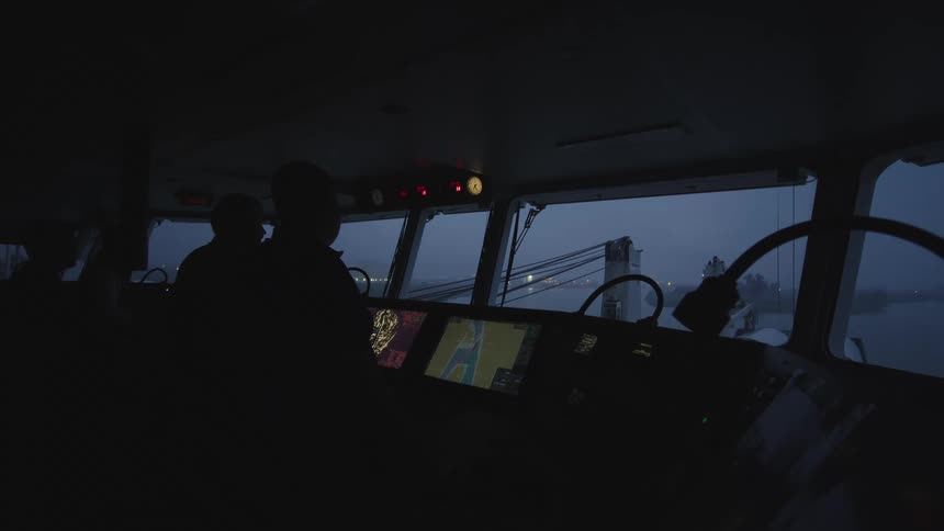 Sailors in silhouette navigate a ship at night from the dark bridge, with glowing screens and a lit bridge visible outside