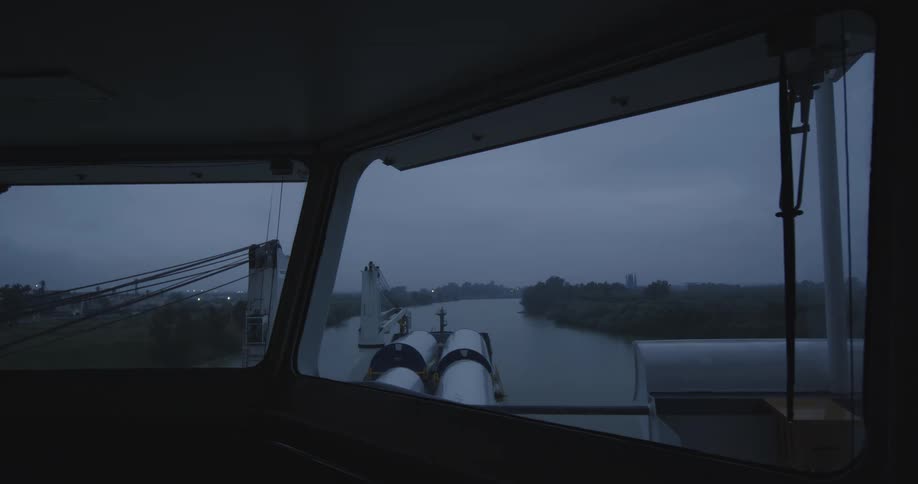 The moody view from inside the bridge of a ship sailing down a river on a dark and overcast evening