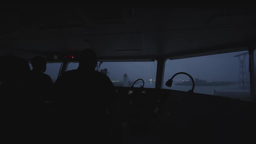 Silhouettes of the ship's crew on the navigation bridge as the vessel moves through an industrial port at dusk