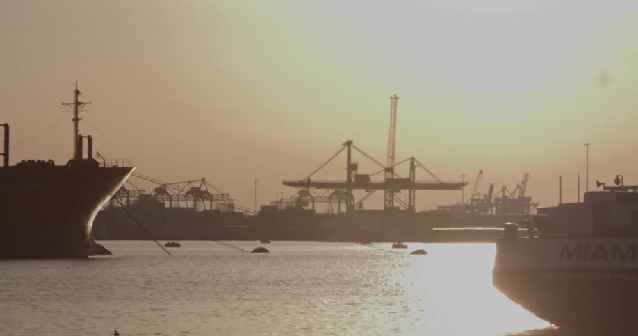Industrial cargo ships and cranes are silhouetted in a hazy harbor during a warm, golden sunset