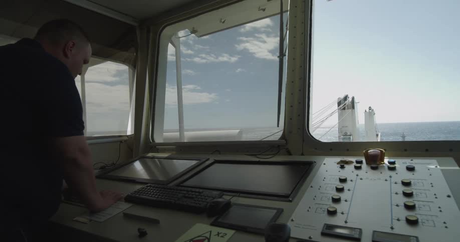 A sailor stands at the control console on the bridge of a large ship sailing on the open ocean