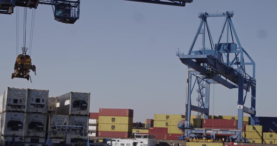 A large gantry crane moves a shipping container at a busy industrial port with stacks of cargo