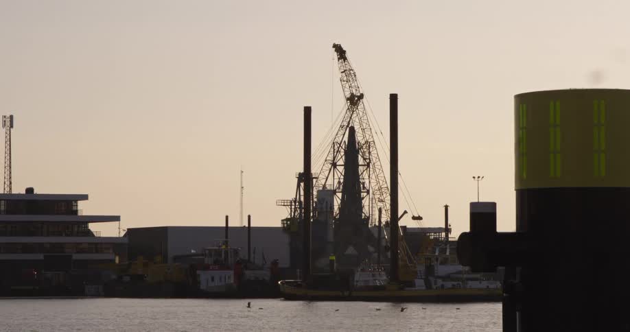 A large construction crane on a floating barge operates in an industrial harbor, silhouetted against the hazy evening sky