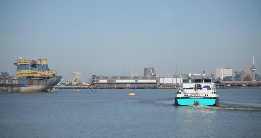 A cargo barge loaded with shipping containers sails through an industrial harbor past a large moored ship on a clear day