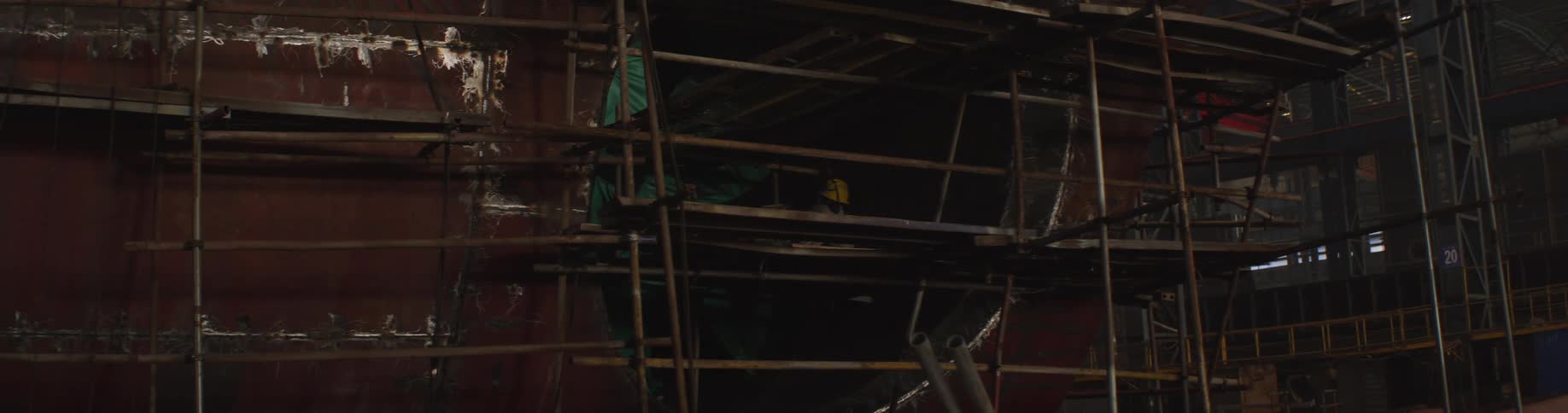 A welder works on the hull of a large ship from scaffolding inside a dark and industrial shipyard