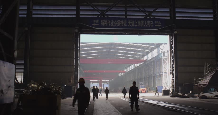 Silhouetted factory workers walk through a massive industrial warehouse towards the light