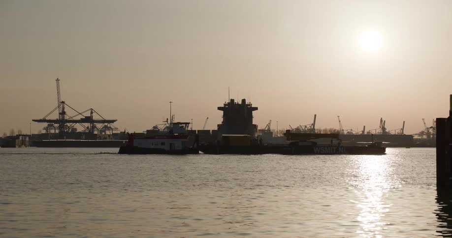 A silhouetted tugboat pushes a barge through a massive industrial harbor at sunset, with numerous cranes lining the background