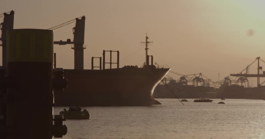 A silhouetted cargo ship sits docked in an industrial harbor at sunset as small boats move across the water