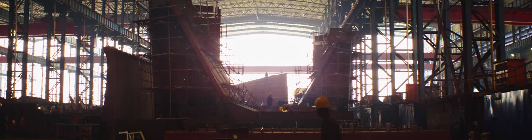 Workers weld the massive hull of a ship under construction inside a vast, sunlit shipyard hangar