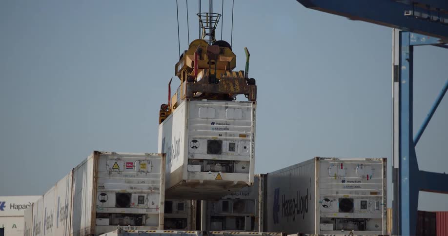 An industrial crane at a shipping port carefully moves and stacks a refrigerated container onto a pile of others