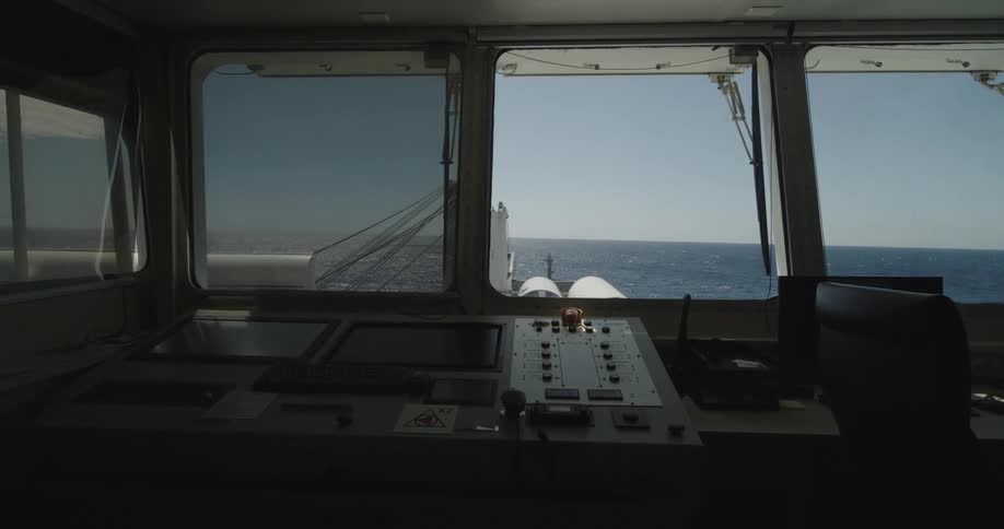 A static shot from the interior of a ship's bridge shows the control panel overlooking the vast blue ocean on a sunny day