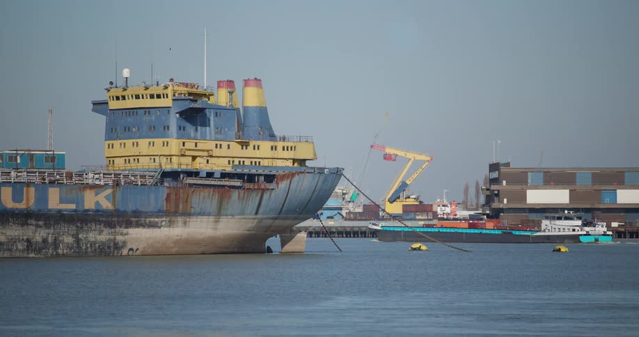 A rusty blue and yellow bulk carrier ship is anchored in a busy industrial harbor as a smaller container barge sails past in the background