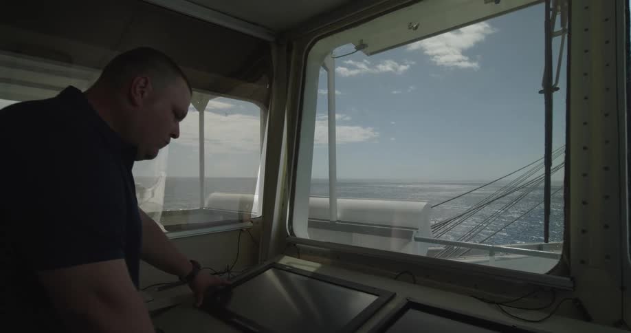 A male captain navigates a large ship from the bridge using a modern touchscreen console with the open ocean visible through the window