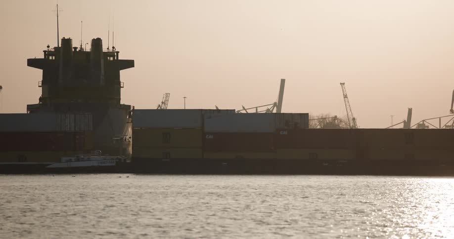 A silhouetted cargo container ship slowly sails through a hazy industrial harbor at sunset