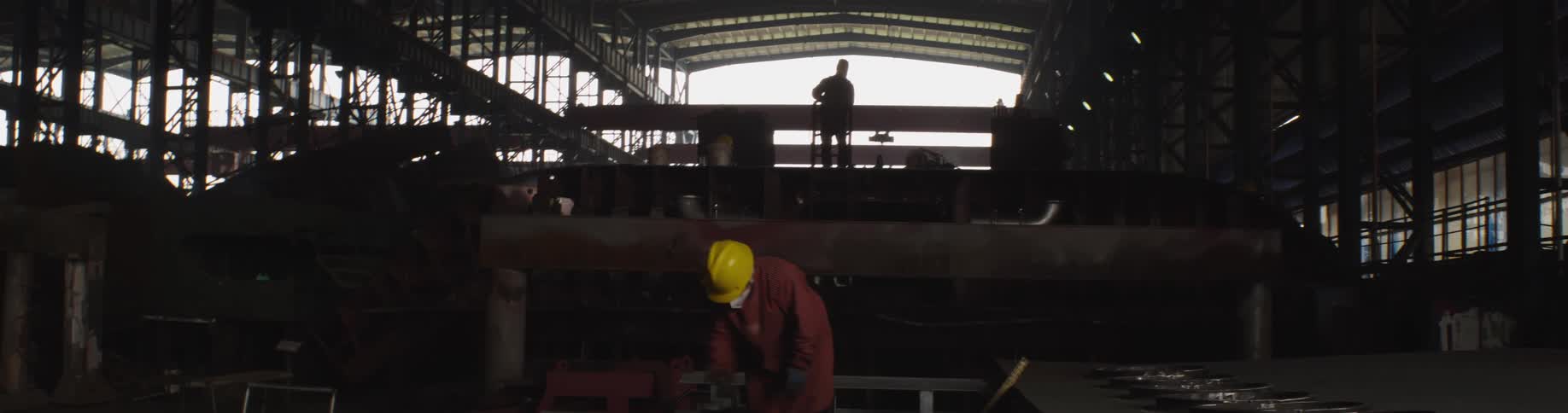 Silhouetted workers toil in a massive industrial hall filled with large metal structures