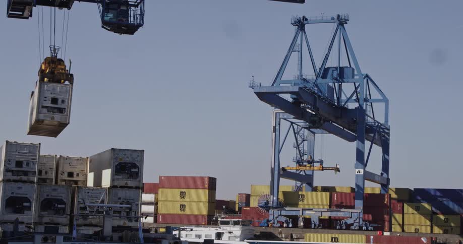 A large blue gantry crane operates at a busy shipping port, moving and stacking colorful cargo containers under a clear sky