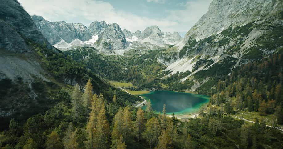 A stunning timelapse captures the transition from lush summer greens to vibrant autumn oranges around a turquoise alpine lake nestled in a majestic mountain valley