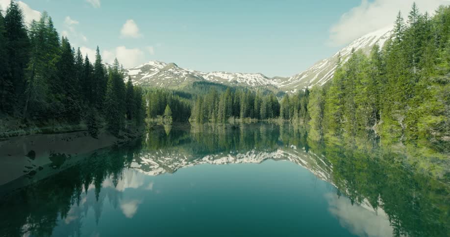 A timelapse captures the transition of seasons from a snowy winter to a lush spring over a tranquil mountain lake reflecting the surrounding forest and peaks
