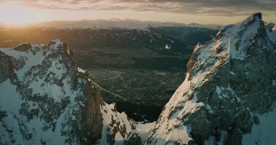 An aerial timelapse shows the sun setting over majestic snowy mountains, revealing the sparkling lights of a city in the valley as day turns to night