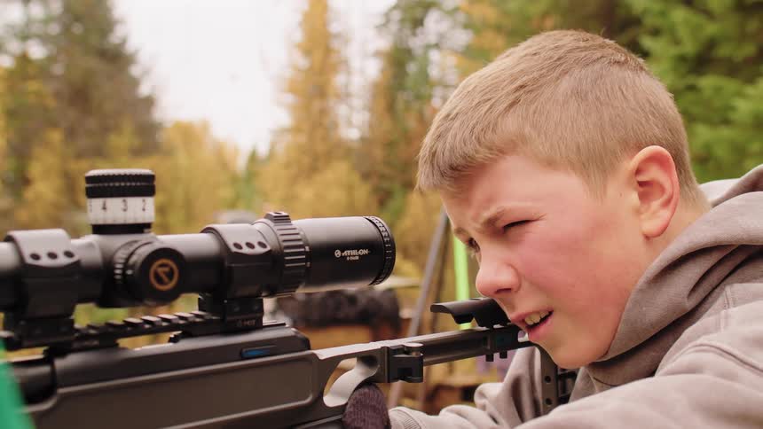 A close-up of a young boy squinting with intense focus as he aims a high-powered rifle through its scope outdoors
