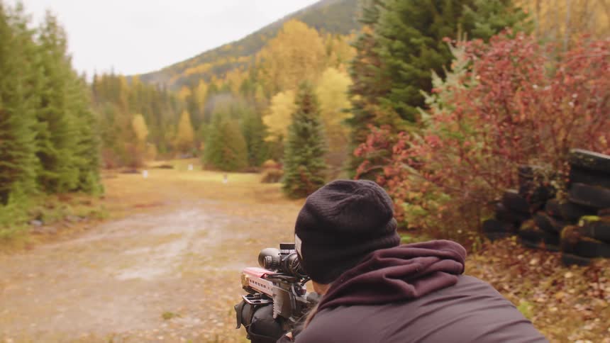 A woman in a black beanie aims a scoped rifle downrange at a target in a beautiful autumn forest setting