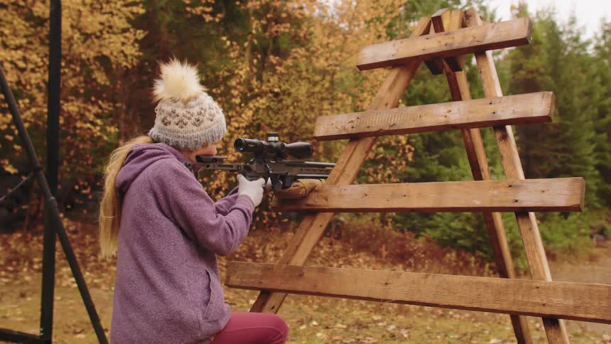 A young girl in a winter hat fires a scoped rifle from a wooden barricade at an outdoor range with her instructor