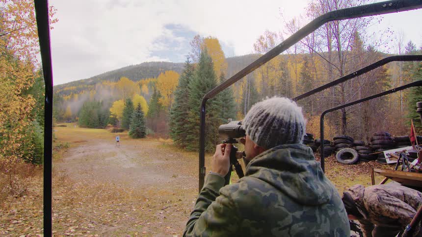 A man in a camo hoodie uses a spotting scope on a tripod at an outdoor shooting range in the autumn forest