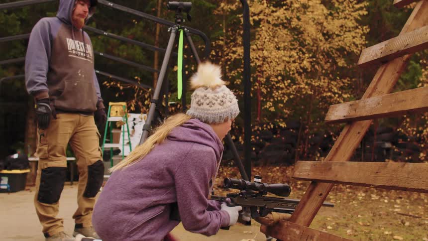 A young woman with a long-range rifle gets into a shooting position on a wooden barricade while her instructor observes