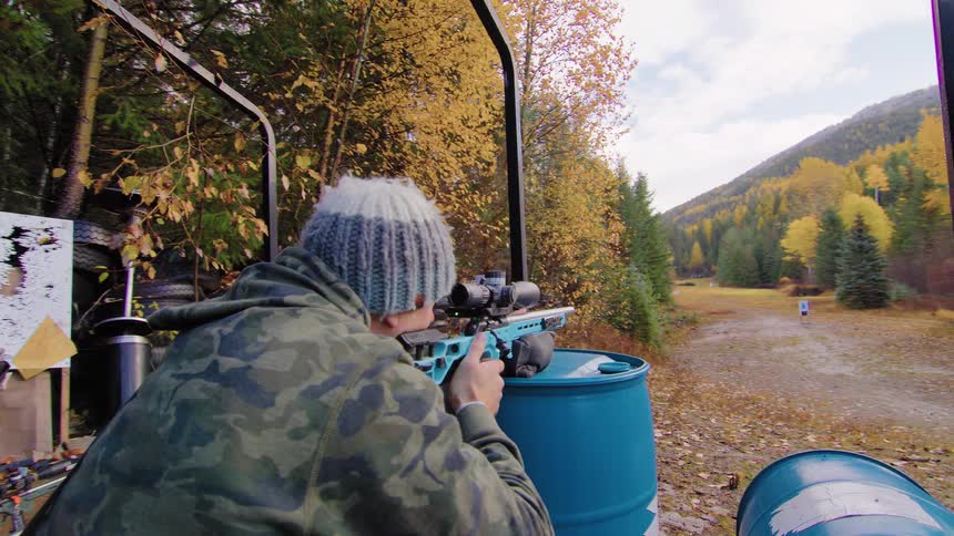 A shooter in camouflage aims a modern blue rifle and fires at a target down an outdoor range in the mountains during autumn