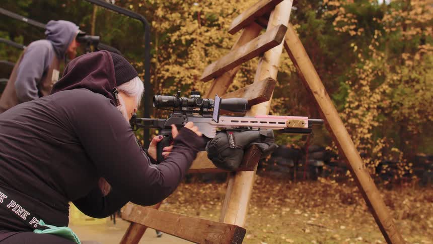 A woman aims and chambers a round in her precision rifle at an outdoor shooting range with a spotter in the background