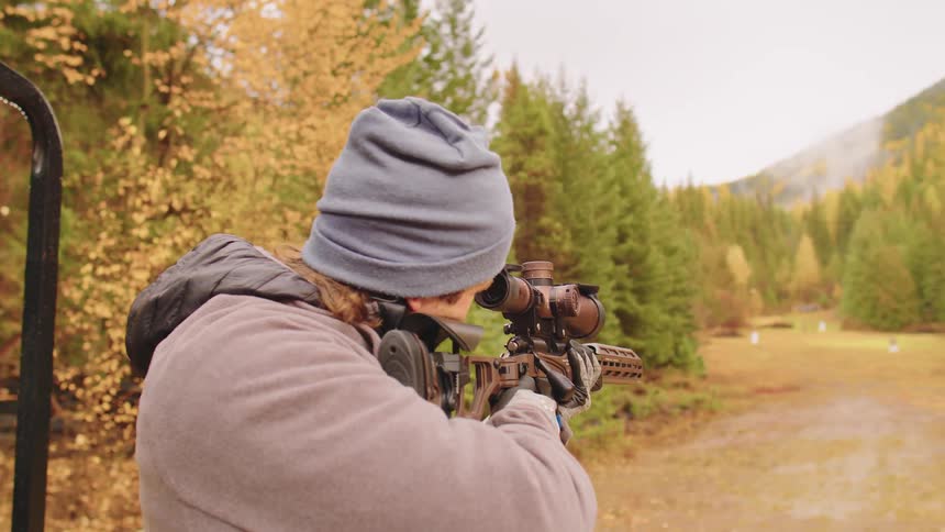 A man in a beanie takes aim and fires a modern scoped rifle in slow motion at an outdoor range surrounded by autumn foliage