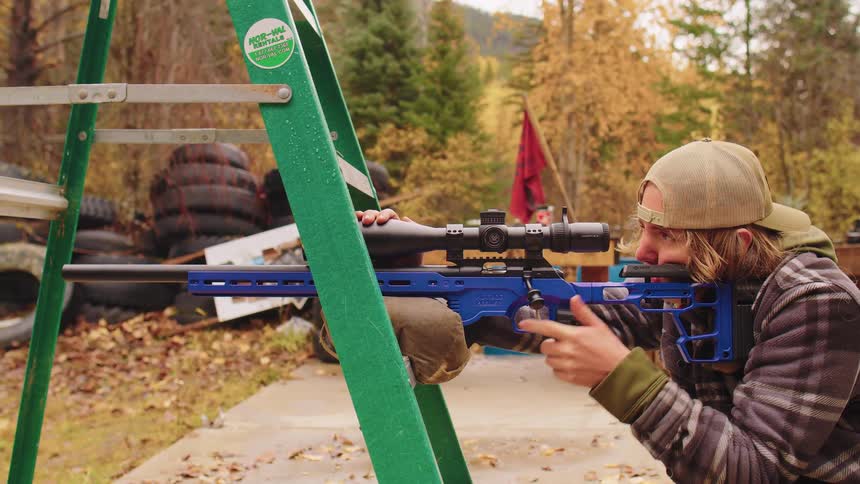 A young man fires a modern bolt-action rifle in slow motion while stabilizing it on a ladder at an outdoor range