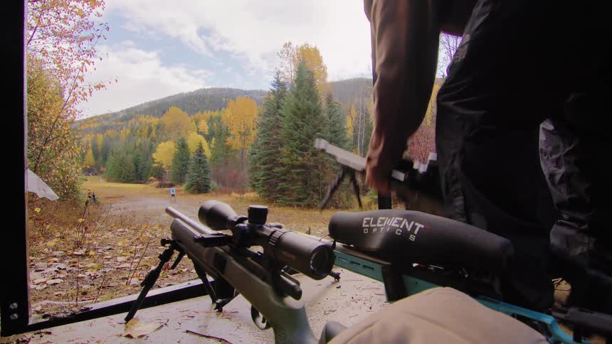 A shooter sets up their rifle at an outdoor range with vibrant autumn foliage in the background