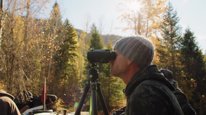 A man in a beanie and camouflage jacket surveys the autumn landscape with binoculars mounted on a tripod on a sunny day