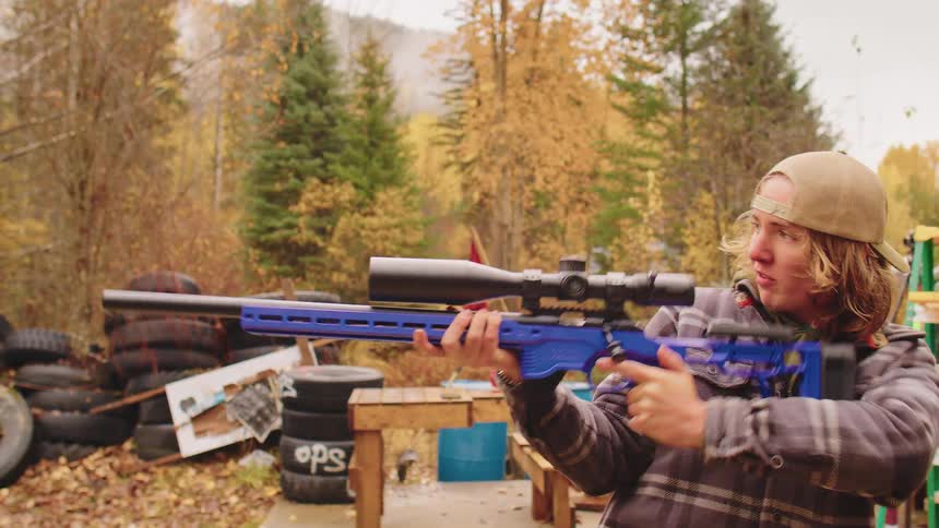 A young man in a plaid jacket aims and cycles the bolt of a modern blue rifle in slow motion in a rustic backyard