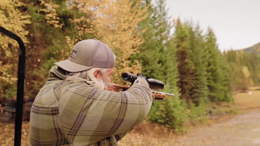 An older man with a long beard aims his scoped rifle at a target down range in an autumn forest