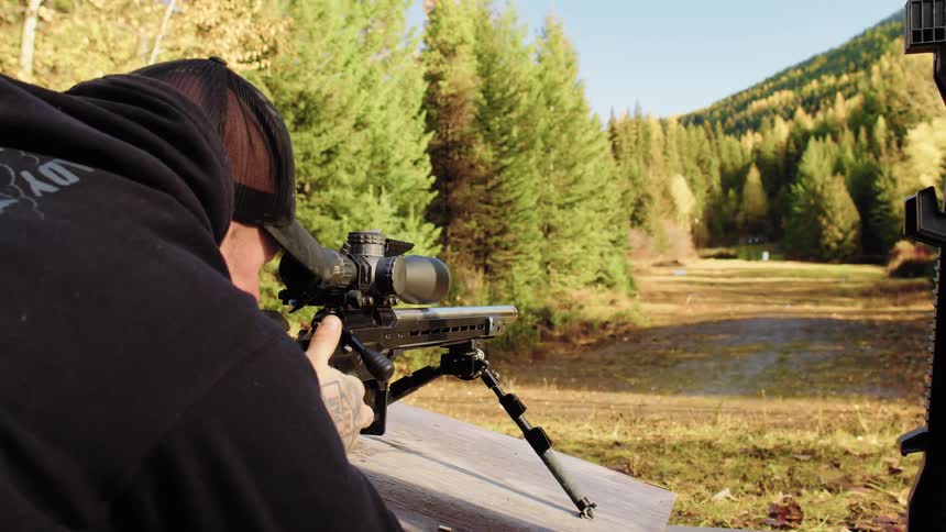 A man at a shooting range aims a long-range rifle, shoots, ejects the spent casing in slow motion, and then picks up the gun