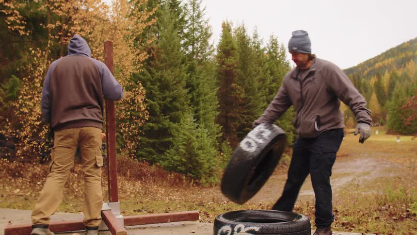 Two men in work clothes set up an outdoor training course with tires and a wooden stand in slow motion