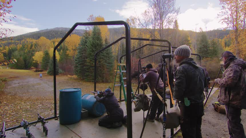 A group of men practice rifle marksmanship at an outdoor shooting range surrounded by autumn foliage