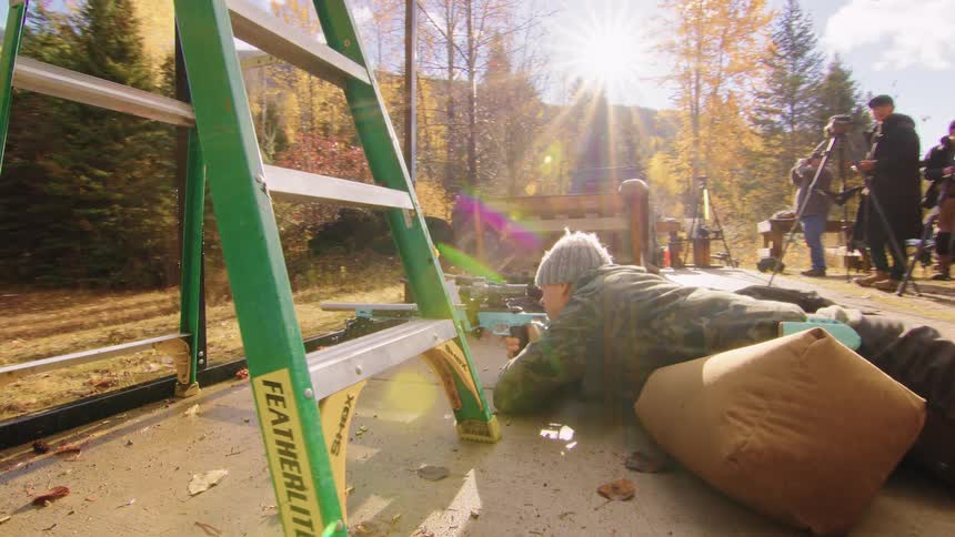A shooter in camouflage gets into a prone position with a rifle under a ladder during an outdoor competition on a sunny autumn day