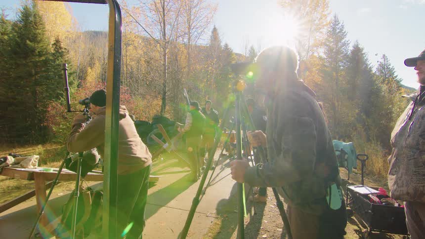 A group of men practices their aim with rifles and spotting scopes at an outdoor shooting range on a sunny autumn day