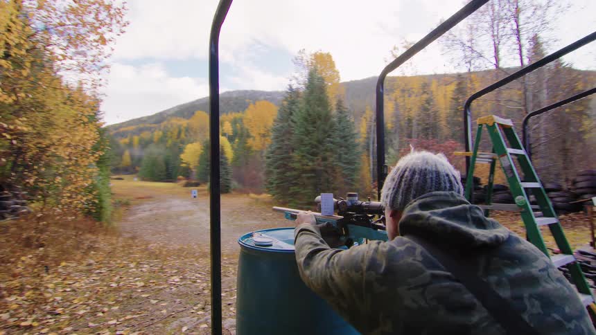 Slow motion shot of a man in a camouflage hoodie steadying his rifle on a barrel for target practice at an outdoor range during autumn