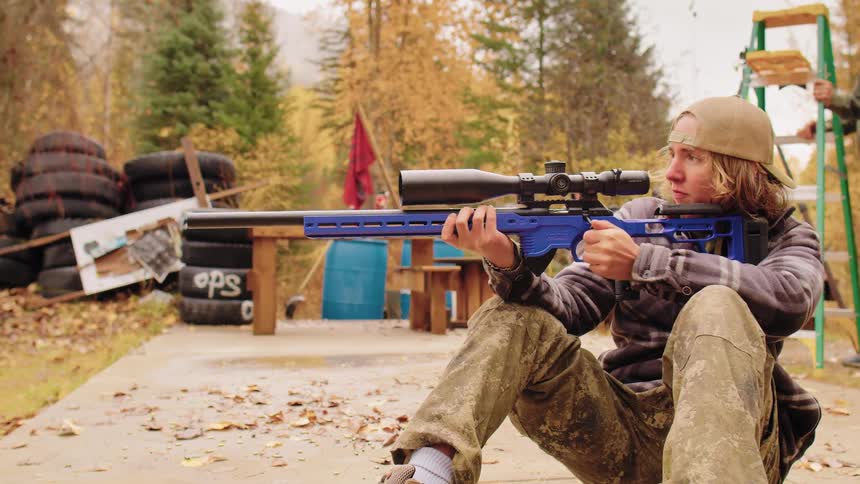 A young man with long hair aims and cocks a blue sniper rifle at an outdoor shooting range during autumn