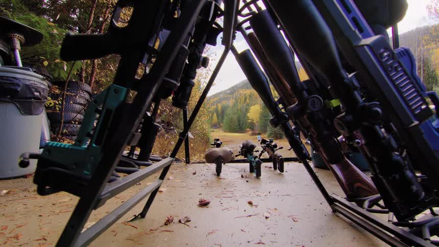 A probe lens pulls back through a gun rack filled with various rifles at an outdoor shooting range in an autumn forest