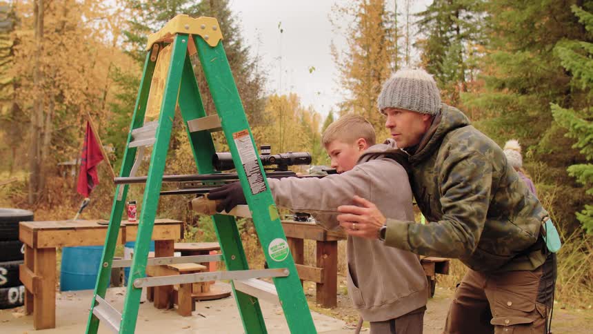 A father teaches his young son how to safely aim and shoot a rifle at an outdoor range in the autumn
