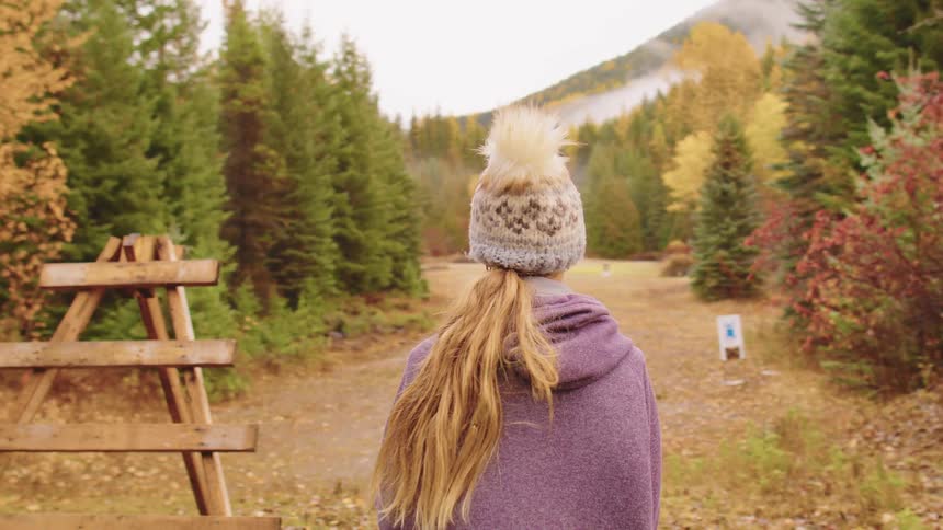 A young woman wearing a warm hat and poncho observes targets through a spotting scope at a rustic shooting range during autumn