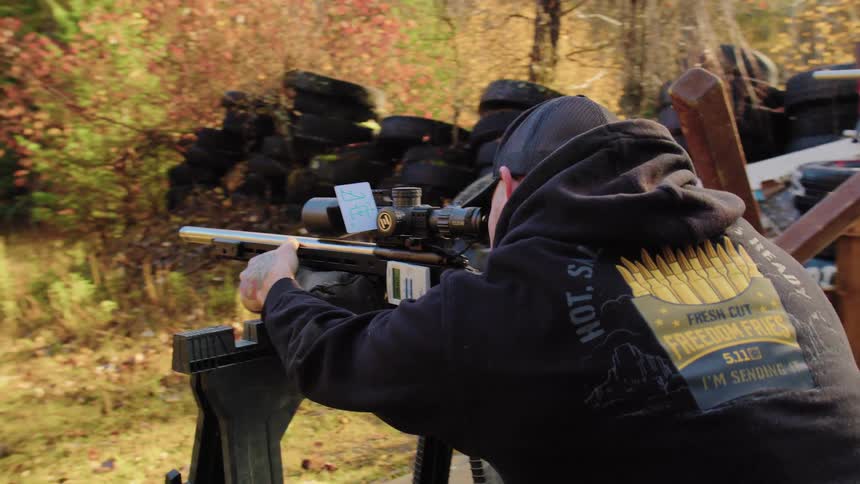 Slow-motion shot follows a man as he aims a long-range scoped rifle down a rural outdoor shooting range during autumn