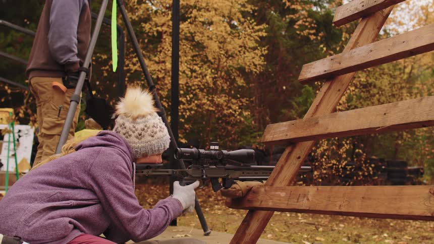 A woman in a beanie carefully aims a scoped rifle from behind a wooden barricade at an outdoor shooting range in autumn